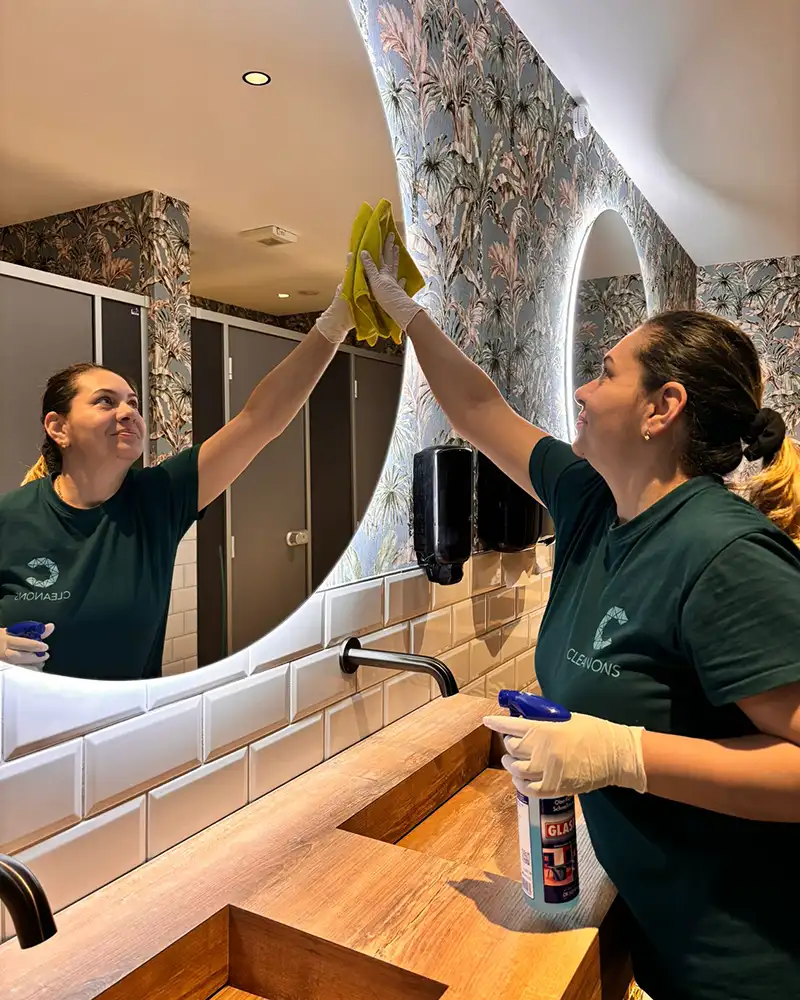Auf dem Foto ist eine Person, die mit Handschuhen an und einem Schwamm in der Hand einen großen runden Spiegel putzt. Der Spiegel hängt neben einem weiteren über zwei hölzernen Waschbecken. Im Spiegel spiegeln sich Toilettenkabinen hinter der Person. Die Person lächelt und trägt ein T-Shirt mit dem Cleanons-Logo.