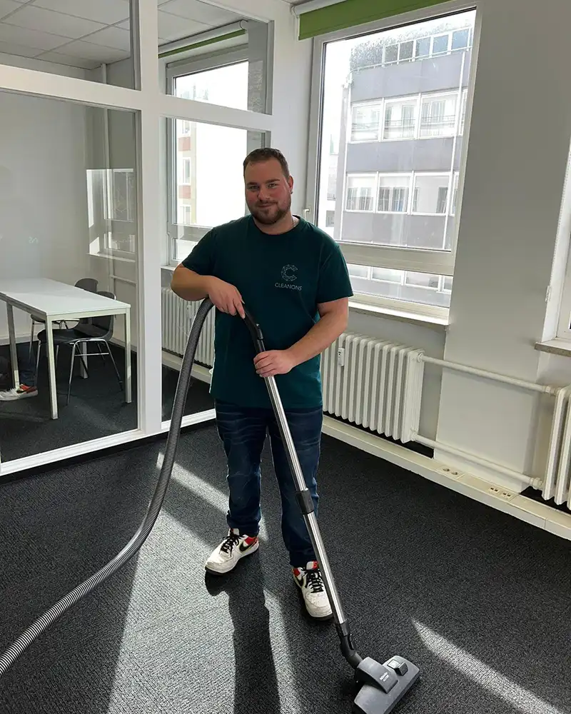 Auf dem Foto ist eine Person beim Staubsaugen eines Teppichbodens. Die Person lächelt freundlich in die Kamera. Auf dem T-Shirt, das sie trägt, ist das Logo von Cleanons. Der Raum ist ein Büro.